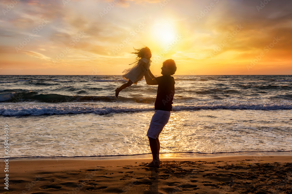 Silhouette of a happy father on vacation holding his daughter in the air on the beach during sunset time