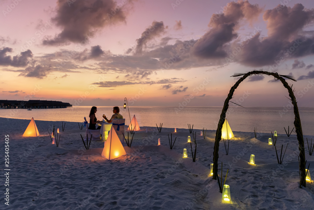 Paar in den Flitterwochen sitzt bei einem privatem Abendessen am Strand und genießt den Sonnenuntergang  - obrazy, fototapety, plakaty 