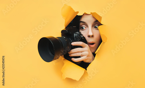 A paparazzi funny girl with a camera looks out from cover and looks at what is happening with her mouth open. Yellow paper, torn hole. Tabloid press. In search of the plot for photo stocks.