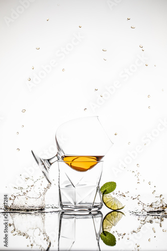 Abstraction splashes and drops with two glasses with whiskey, lime, and mint on the mirror white background. Food in motion