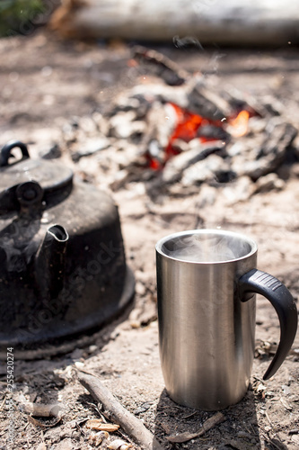 Cup with a hot drink and a kettle stand on the ground, against a blurred background of a bonfire with embers, on a sunny morning in the camp.