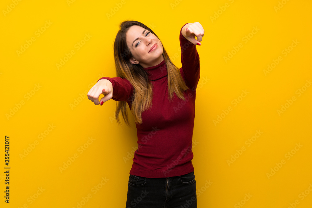Woman with turtleneck over yellow wall points finger at you while smiling Woman with turtleneck over yellow wall points finger at you while smiling