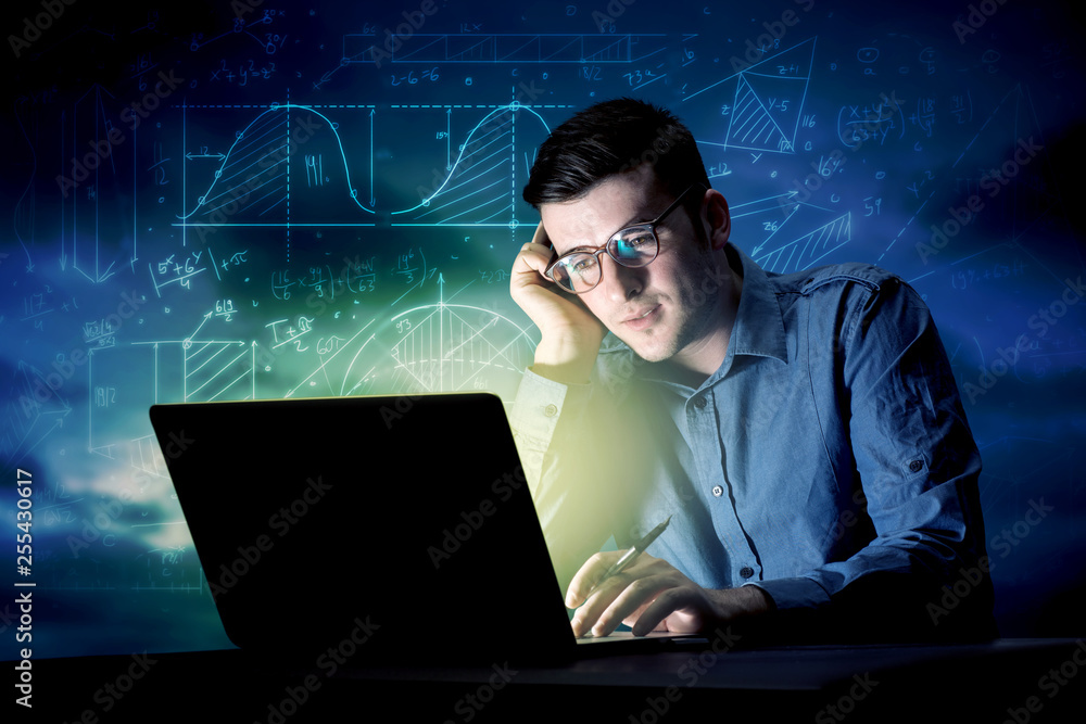 Young handsome businessman working late at night in the office with blue mathematic charts in the background Young handsome businessman working late at night in the office with blue mathematic charts in the background
