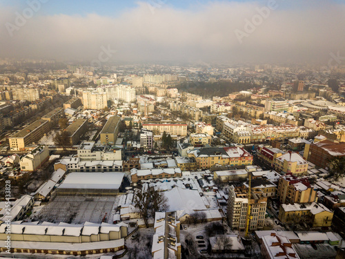 Aerial black and white winter top view of modern city center with tall buildings and parked cars on snowy streets.