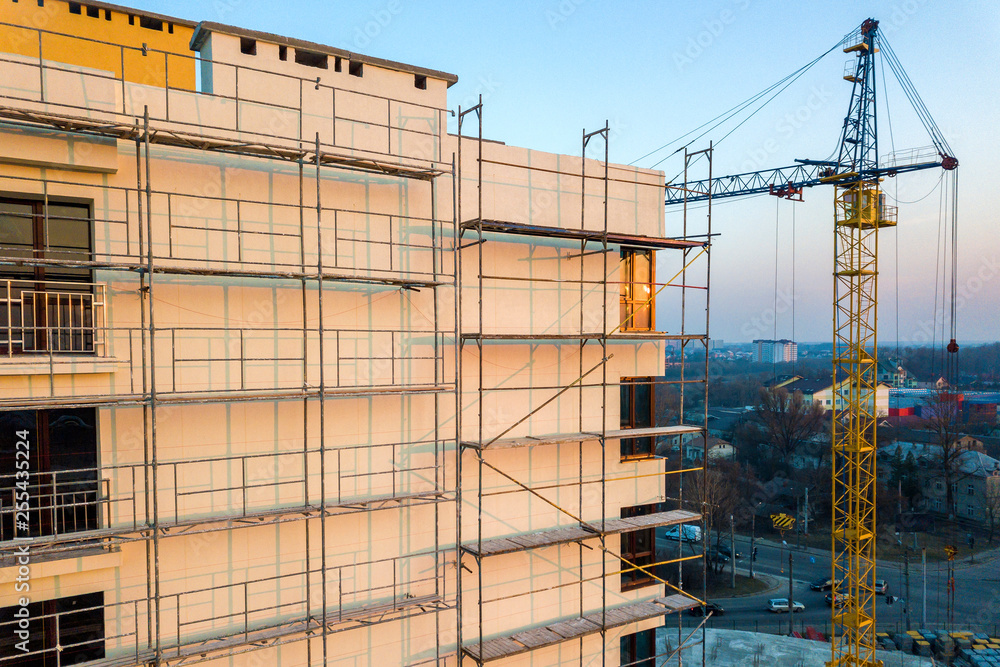 Apartment or office tall unfinished building under construction. Brick wall in scaffolding, shiny windows and tower crane on urban landscape and blue sky background. Drone aerial photography. Apartment or office tall unfinished building under construction. Brick wall in scaffolding, shiny windows and tower crane on urban landscape and blue sky background. Drone aerial photography.