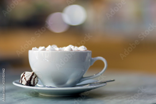 Mug of coffee with marshmallows on porcelain plate on blurred colorful interior bokeh background.