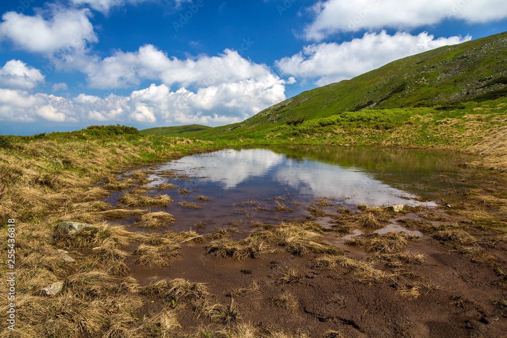 Blue sky with bright white clouds reflected in small lake between green hills on sunny day. Summer panorama.
