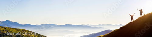 Small dark silhouettes of tourist travelers on steep mountain slope at sunrise on copy space background of valley covered with white puffy clouds and bright clear sky.