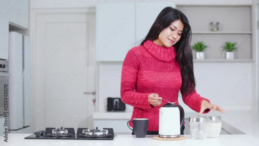 Pretty young woman making hot tea and enjoying it while wearing winter clothes in the kitchen at home. Shot in 4k resolution Pretty young woman making hot tea and enjoying it while wearing winter clothes in the kitchen at home. Shot in 4k resolution