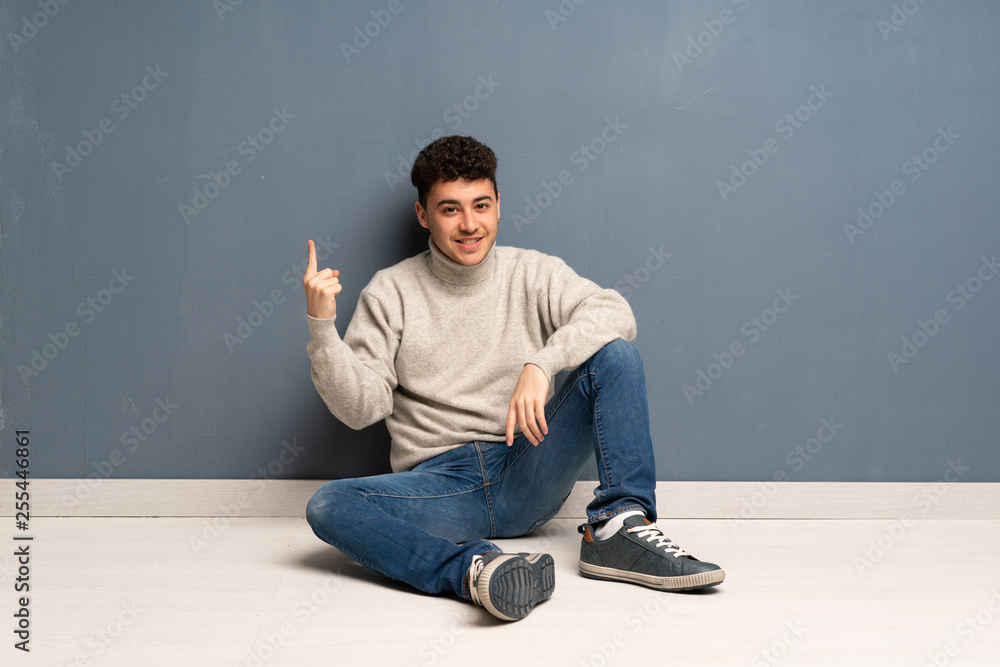 Young man sitting on the floor showing and lifting a finger in sign of the best Young man sitting on the floor showing and lifting a finger in sign of the best