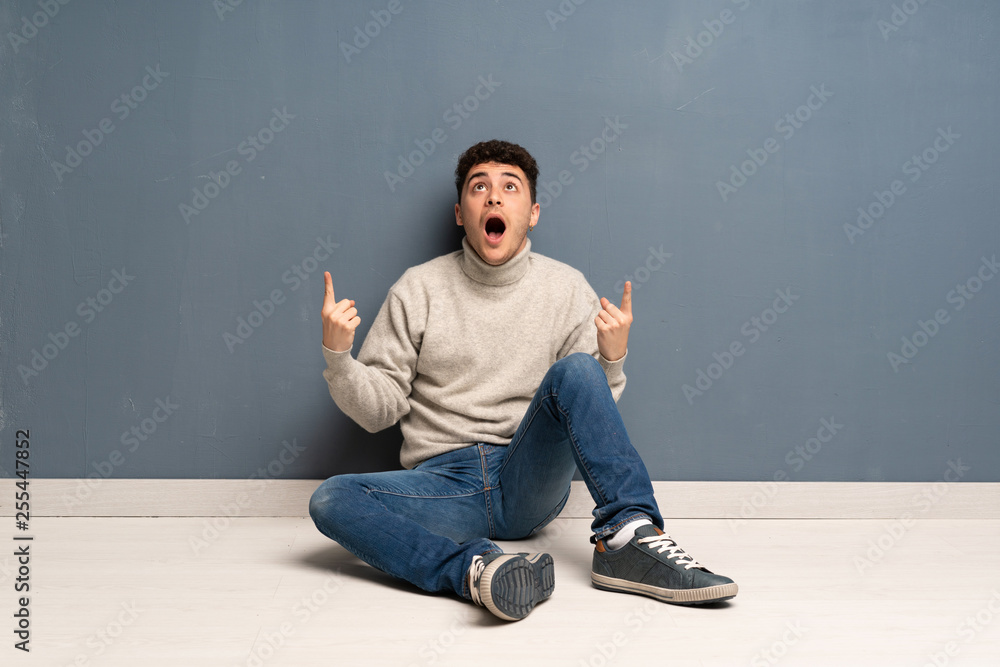 Young man sitting on the floor surprised and pointing up Young man sitting on the floor surprised and pointing up