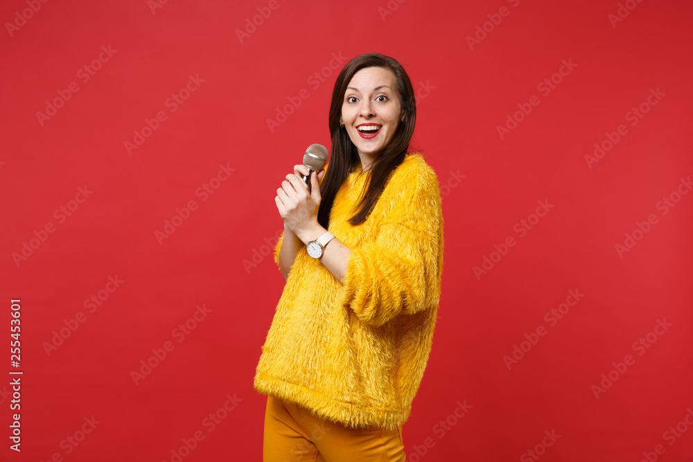 Portrait of excited young woman in yellow fur sweater sing song, holding microphone isolated on bright red wall background in studio. People sincere emotions, lifestyle concept. Mock up copy space. Portrait of excited young woman in yellow fur sweater sing song, holding microphone isolated on bright red wall background in studio. People sincere emotions, lifestyle concept. Mock up copy space.