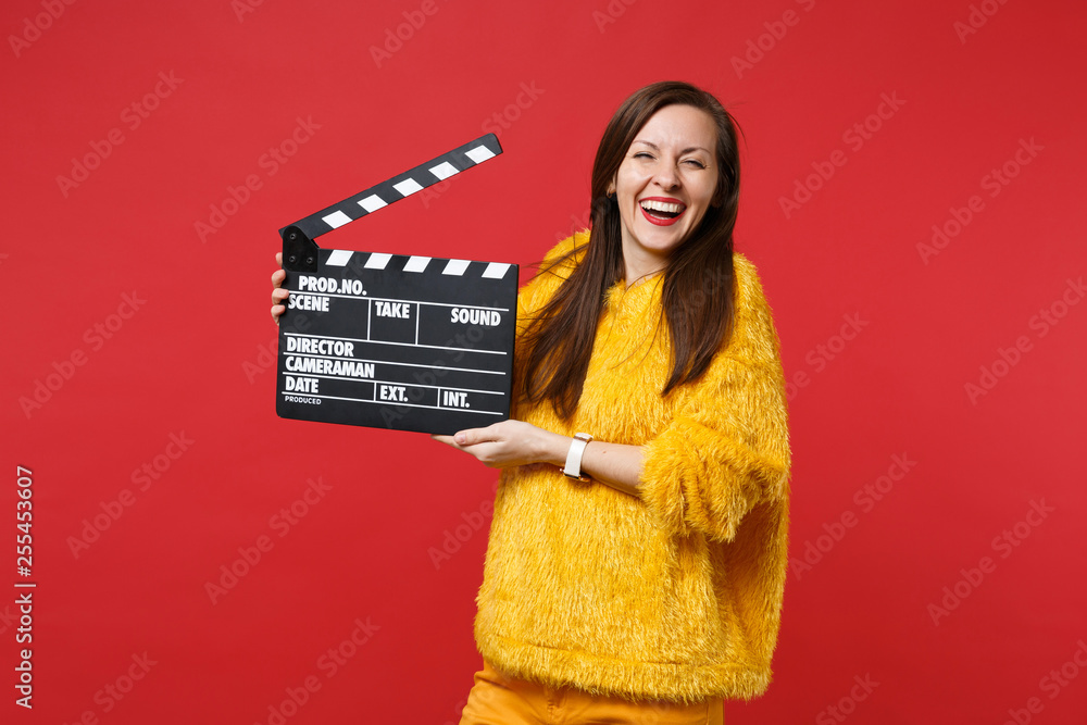 Portrait of laughing young woman in yellow fur sweater holding classic black film making clapperboard isolated on bright red background. People sincere emotions, lifestyle concept. Mock up copy space.