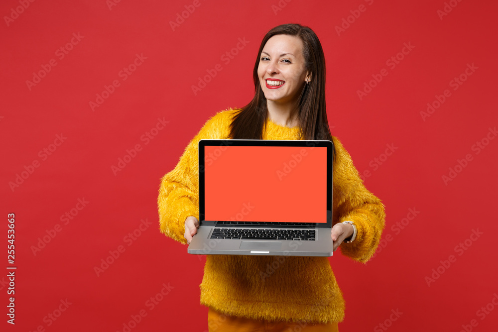 Charming young woman in yellow fur sweater holding laptop pc computer with blank empty screen isolated on red wall background in studio. People sincere emotions, lifestyle concept. Mock up copy space. Charming young woman in yellow fur sweater holding laptop pc computer with blank empty screen isolated on red wall background in studio. People sincere emotions, lifestyle concept. Mock up copy space.