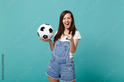 Laughing woman football fan cheer up support favorite team with soccer ball pointing index finger on camera isolated on blue turquoise wall background. People emotions, sport family leisure concept. © ViDi Studio