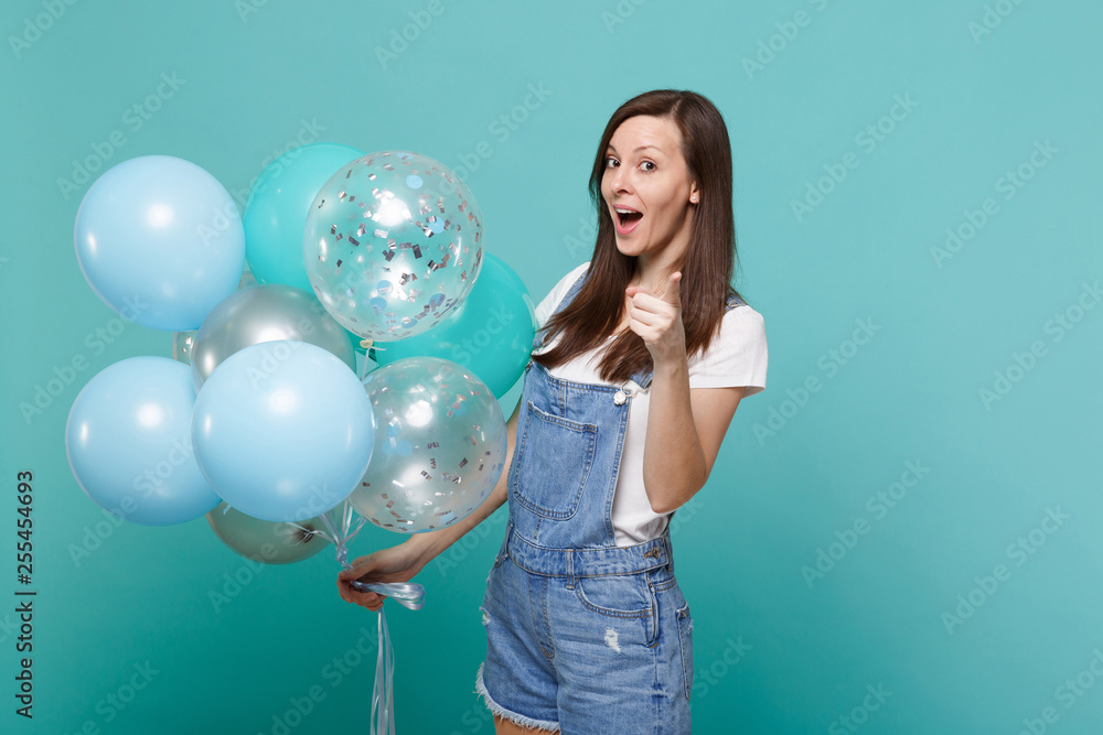 Funny young woman in denim clothes pointing index finger on camera, celebrating, hold colorful air balloons isolated on blue turquoise wall background. Birthday holiday party, people emotions concept. Funny young woman in denim clothes pointing index finger on camera, celebrating, hold colorful air balloons isolated on blue turquoise wall background. Birthday holiday party, people emotions concept.