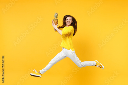 Smiling young woman in casual clothes jumping, hold fresh ripe pineapple fruit isolated on yellow orange wall background in studio. People vivid lifestyle, relax vacation concept. Mock up copy space.