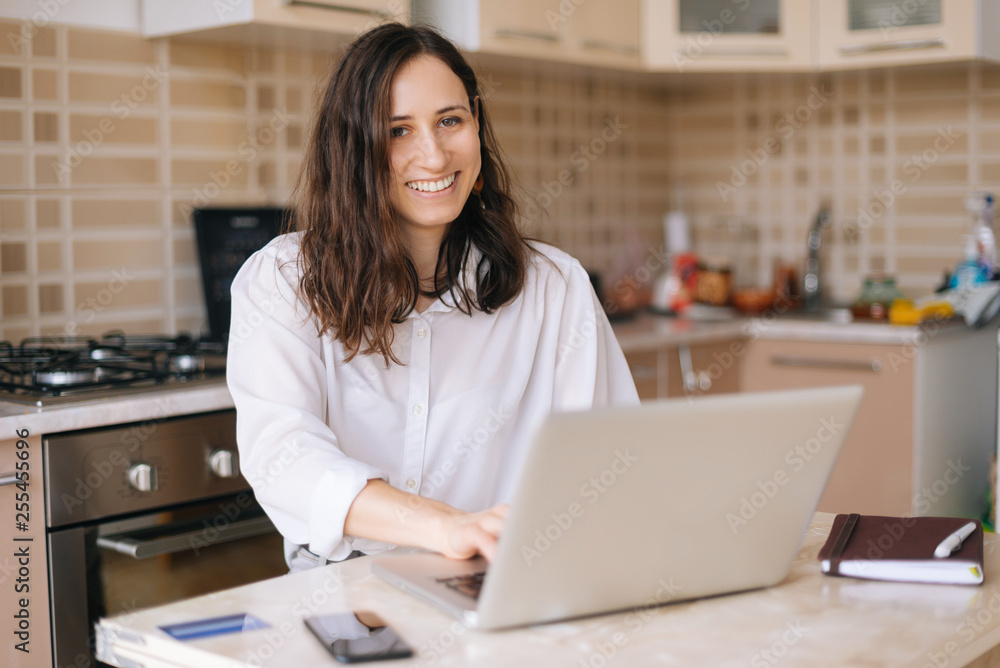 Smiling young woman using laptopand looking at camera in the kitchen at home Smiling young woman using laptopand looking at camera in the kitchen at home