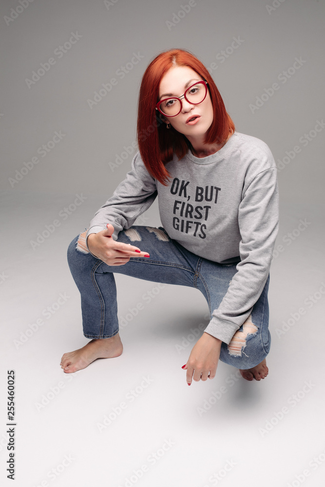 Swag and confident red haired girl in jeans and sweatshirt posing at gray  studio, looking at