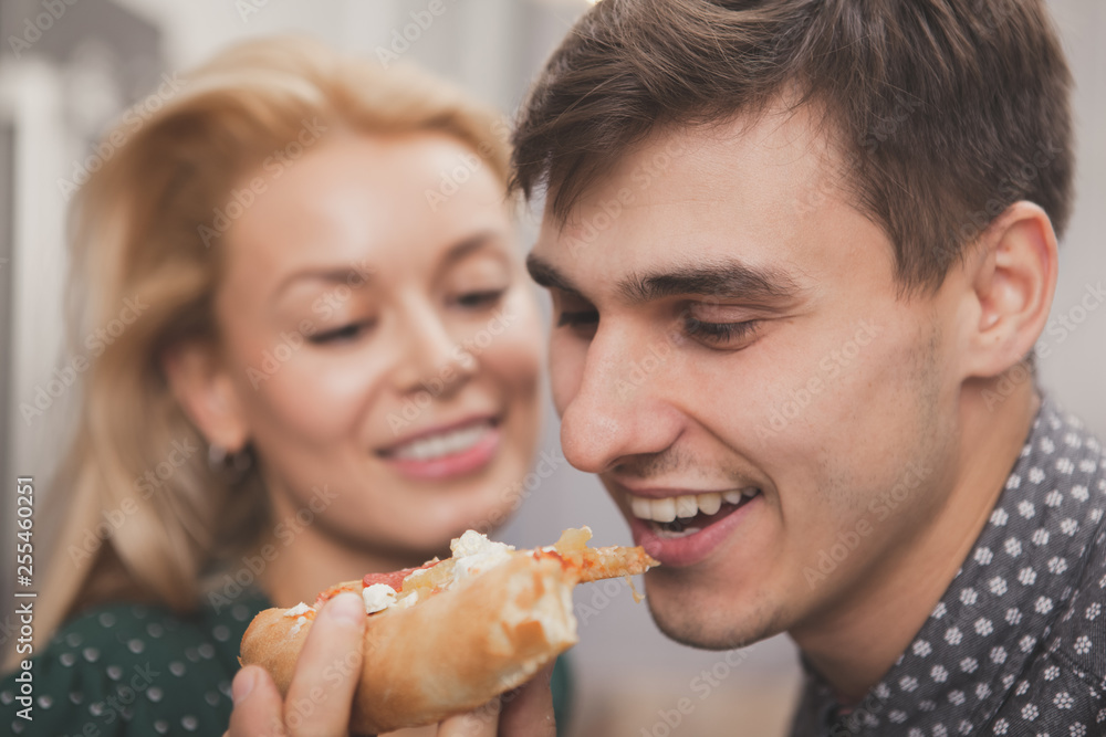 Close up of a young cheerful handsome man biting piece of pizza, his beautiful girlfriend is holding. Happy couple enjoying dining at home, ordering pizza. Food, hunger, meal concept Close up of a young cheerful handsome man biting piece of pizza, his beautiful girlfriend is holding. Happy couple enjoying dining at home, ordering pizza. Food, hunger, meal concept