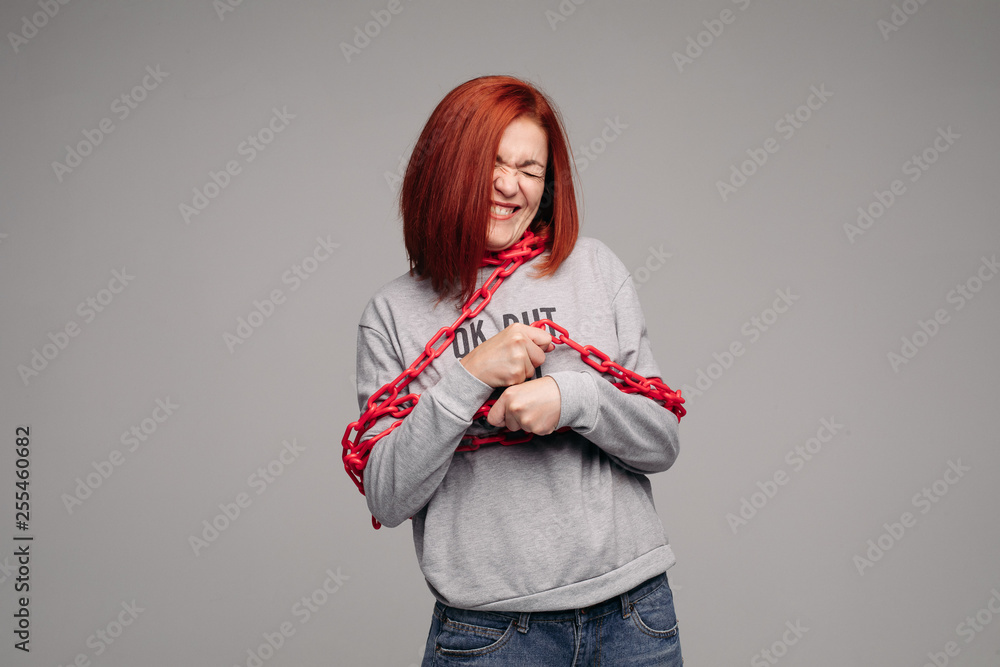 Studio portrait of a girl entangled in a chain. A red-haired woman with brown hair tries to get rid of the chain. The concept of a life-like situation. Isolated on a gray background. Studio portrait of a girl entangled in a chain. A red-haired woman with brown hair tries to get rid of the chain. The concept of a life-like situation. Isolated on a gray background.