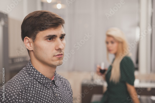 Handsome man looking away thoughtfully, his wife drinking wine on the background. Depressed man looking upset after quarreling with his wife. Couple fighting at home. Depression, divorce concept