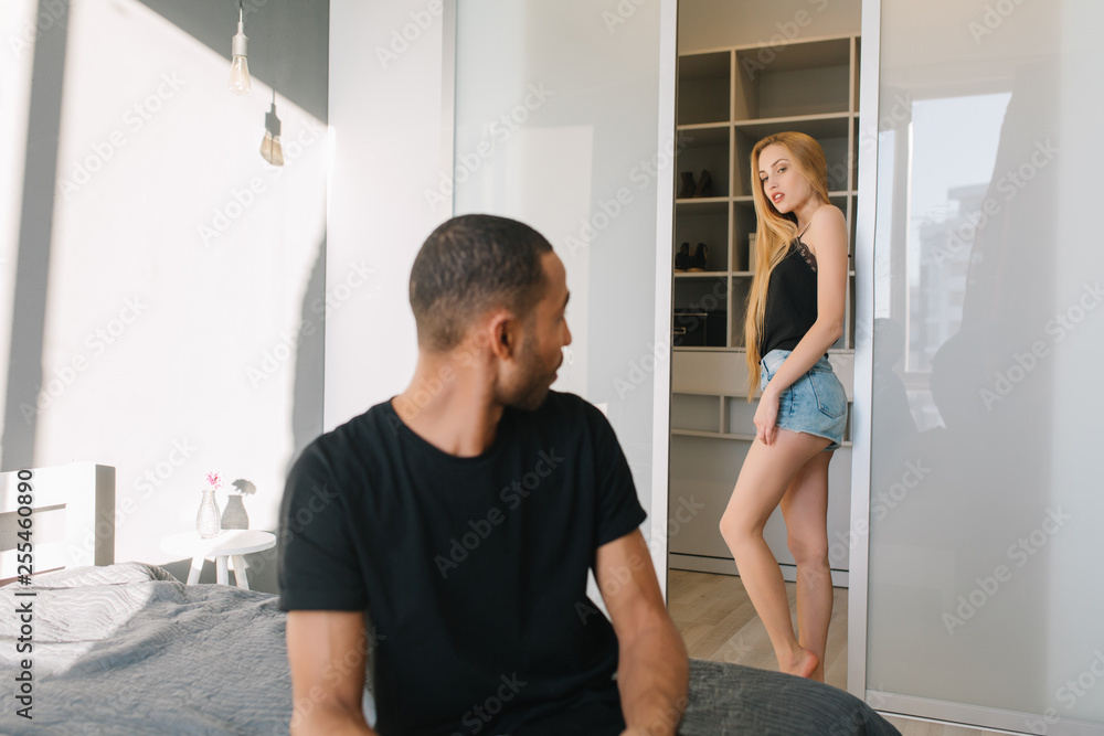 Fototapeta premium Attractive young woman with long blonde hair in shorts behind handsome guy on bed looking at her. Chilling at home, lovely couple, having fun, relationship, leisure, weekends