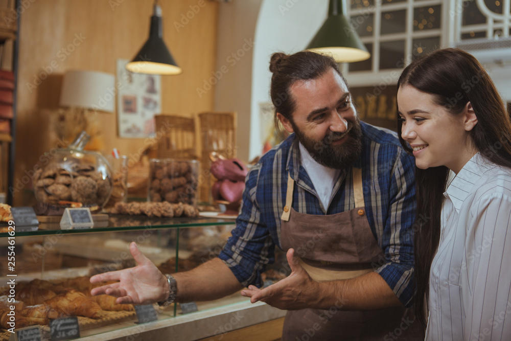 Friendly mature male baker enjoying working at his bakery shop, talking to a customer pointing at the display full of freshly baked pastry. Lovely woman buying desserts at bakery, copy space Friendly mature male baker enjoying working at his bakery shop, talking to a customer pointing at the display full of freshly baked pastry. Lovely woman buying desserts at bakery, copy space