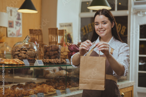 Beautiful cheerful female baker smiling, holding out paper shopping bag to the camera, working at her cafe. Lovely female confectioner selling delicious food at the bakery, copy space