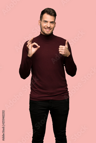 Man with turtleneck sweater showing ok sign with and giving a thumb up gesture over pink background © luismolinero