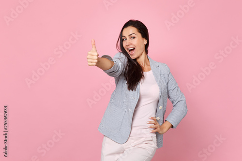 Portrait of joyful laughing young woman in striped jacket standing  showing thumb up isolated on pink pastel wall background in studio. People sincere emotions  lifestyle concept. Mock up copy space.
