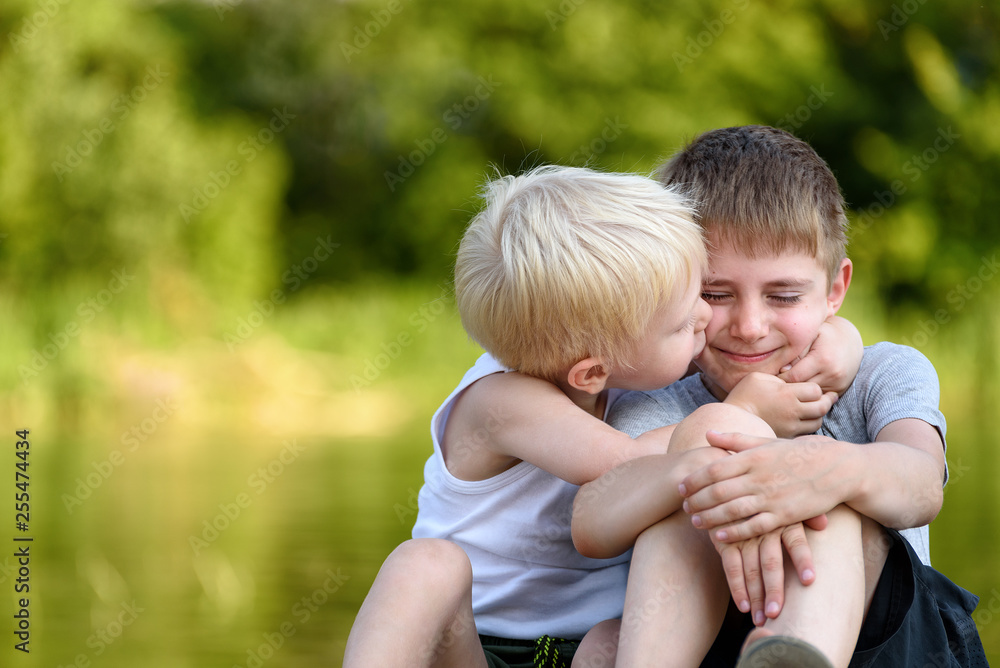 Two little brothers are sitting outdoors. One kisses the other on the cheek. Blurred green trees in the distance. Concept of friendship and fraternity. Two little brothers are sitting outdoors. One kisses the other on the cheek. Blurred green trees in the distance. Concept of friendship and fraternity.