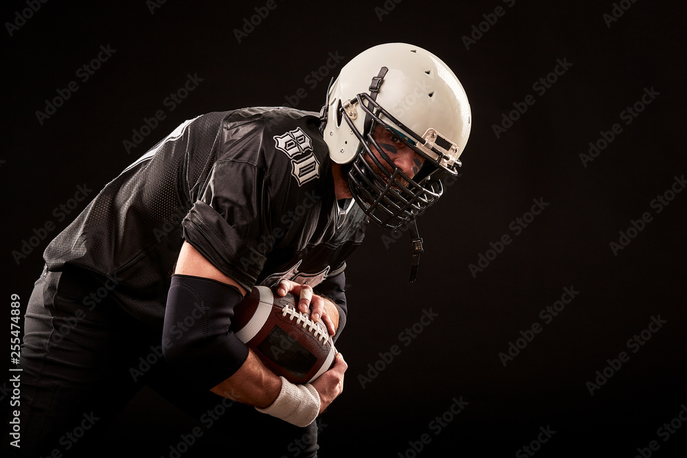 American football player in dark uniform with the ball is preparing to attack on a black background. American football player in dark uniform with the ball is preparing to attack on a black background.