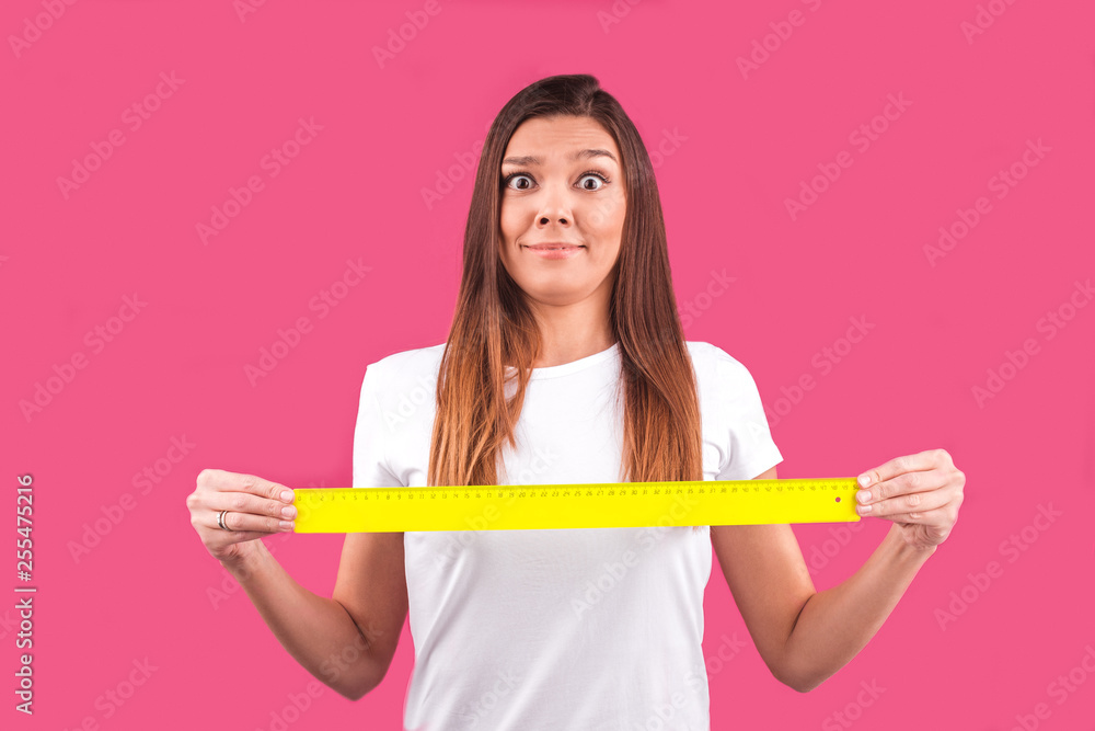 a young surprised brunette girl holds a plastic yellow ruler in front of her Breasts on a pink background with both hands a young surprised brunette girl holds a plastic yellow ruler in front of her Breasts on a pink background with both hands