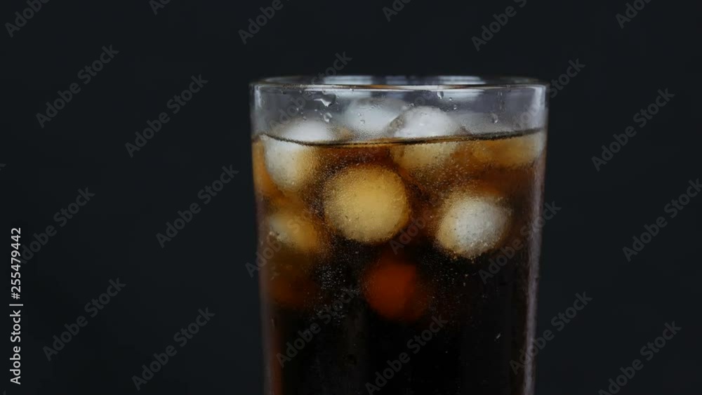 Bubbles of cold cola in a transparent long glass with ice rotating on a black background close up view Bubbles of cold cola in a transparent long glass with ice rotating on a black background close up view