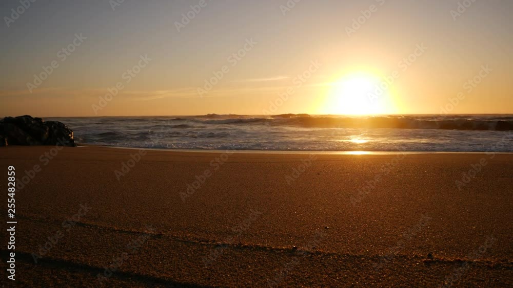Low angle, wide view over sandy beach at sunset with sun on horizon and boulder on left Low angle, wide view over sandy beach at sunset with sun on horizon and boulder on left