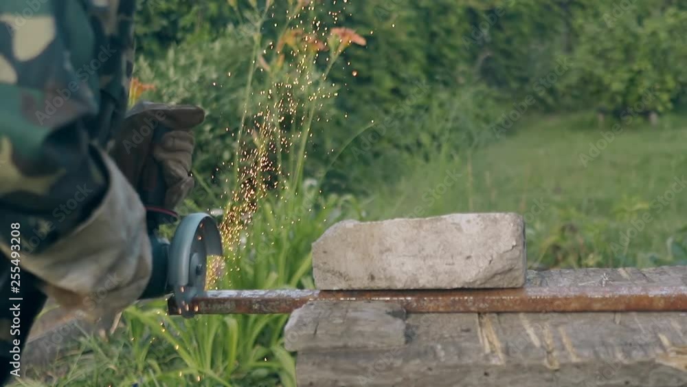 man in work wear cuts long metal part with disc saw on table against grass and flowers slow motion backside view man in work wear cuts long metal part with disc saw on table against grass and flowers slow motion backside view