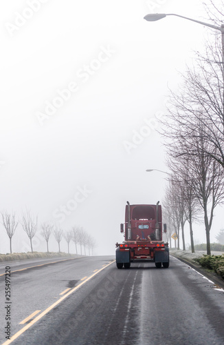 Dark red big rig semi truck tractor moving ahead on foggy winter road with trees on the sides