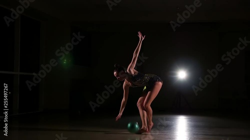 Slim cute attractive girl athlete in bright colorful swimsuit performs elements of rhythmic gymnastics with ball in dark gym with beautiful staged light and rear light