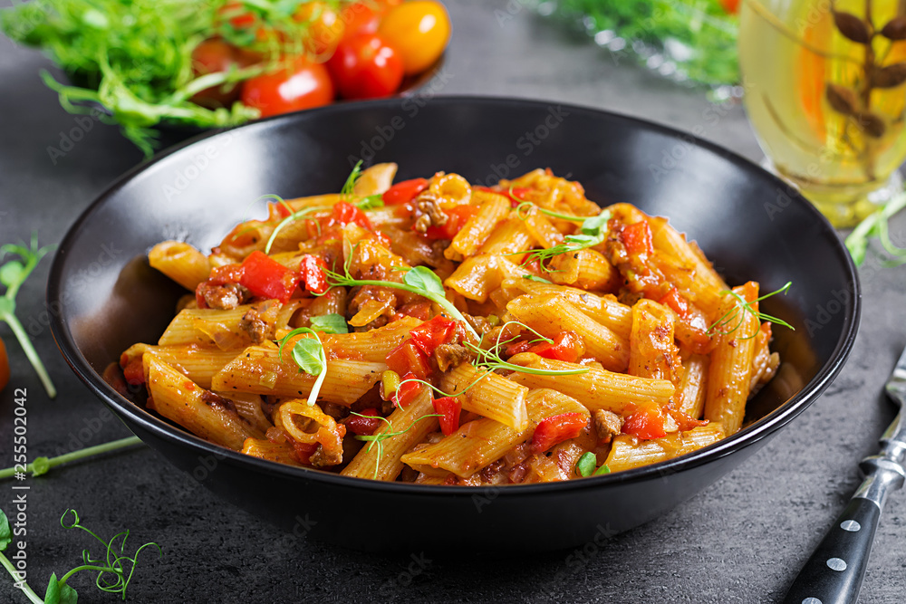 Penne pasta in tomato sauce with meat, tomatoes decorated with pea sprouts on a dark table.