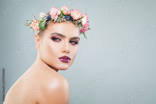 Beautiful woman with colorful makeup in wreath of flowers and leaves on light blue background with copy space
