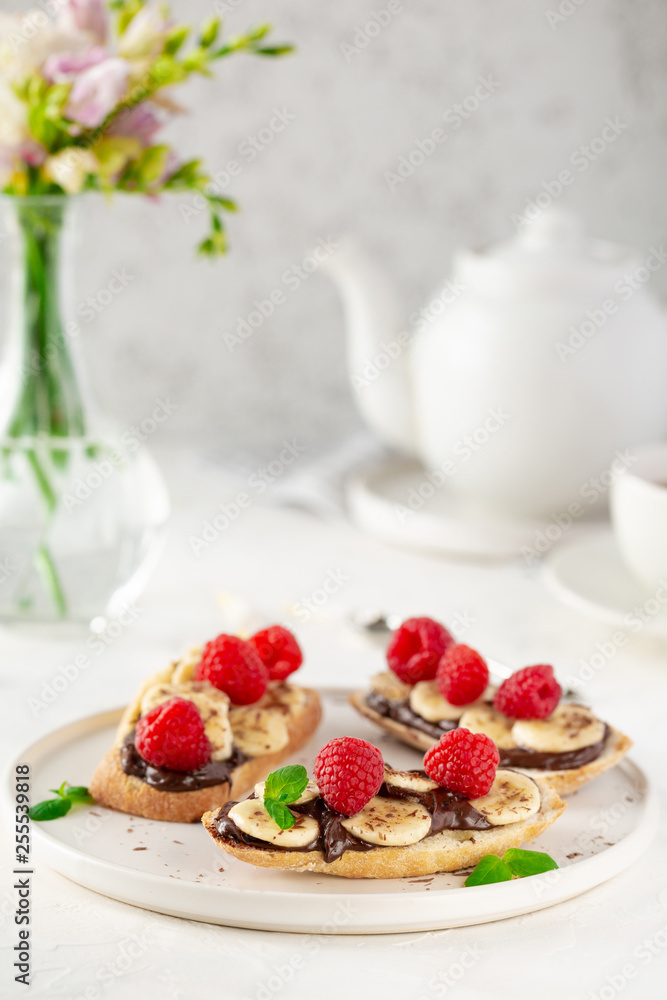 Front view of toast bread with topping - chocolate paste, bananas, raspberries and mint on white background with flowers and teapot. Tasty sweet summer breakfast with toasts and cup of tea Front view of toast bread with topping - chocolate paste, bananas, raspberries and mint on white background with flowers and teapot. Tasty sweet summer breakfast with toasts and cup of tea