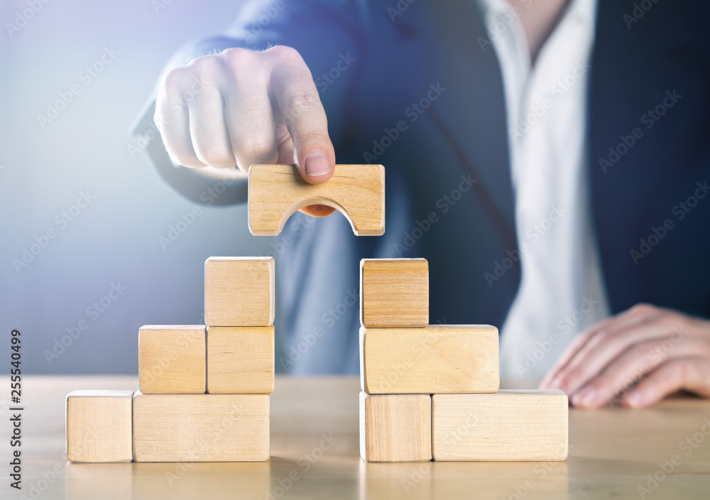 Business man bridging the gap between two towers or parties made from wooden blocks; conflict management or mediator concept Business man bridging the gap between two towers or parties made from wooden blocks; conflict management or mediator concept