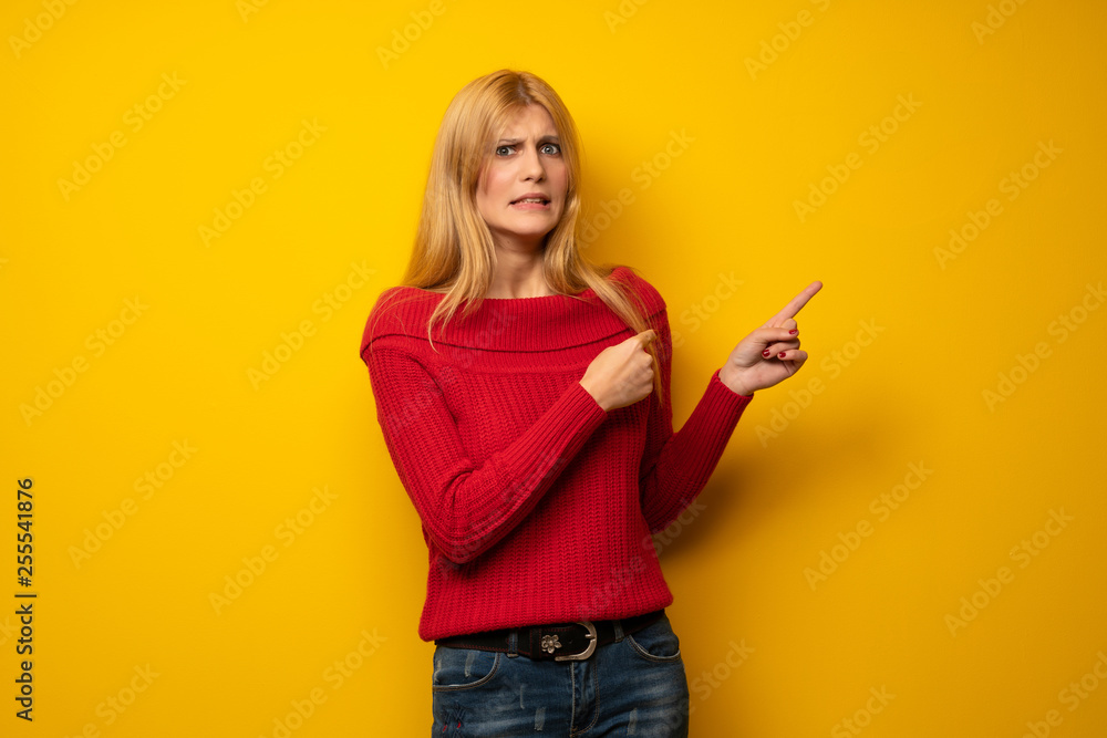 Blonde woman over yellow wall frightened and pointing to the side Blonde woman over yellow wall frightened and pointing to the side