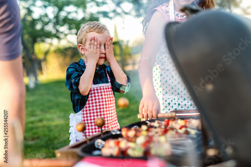 Little chef with apron holding hands on eyes while standing next to grill in backyard. Family gathering concept.