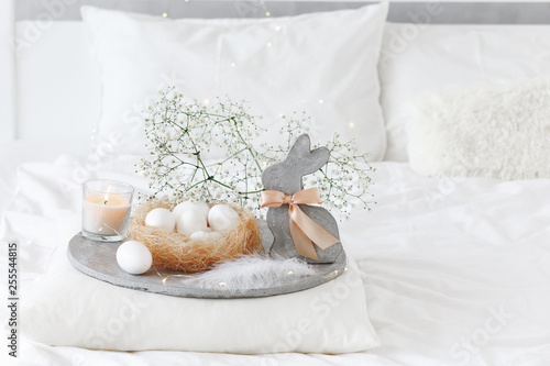 White modern bedroom with Easter decoration. Bed with white bedding set, pillows, concrete tray, nest with white eggs, decorative bunny figure, candle and gypsophila flowers.