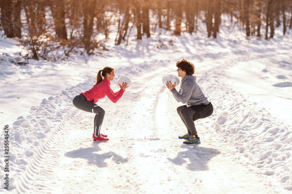 Sporty couple doing squats with big snowballs in hands. Wintertime, healthy lifestyle concept. Sporty couple doing squats with big snowballs in hands. Wintertime, healthy lifestyle concept.