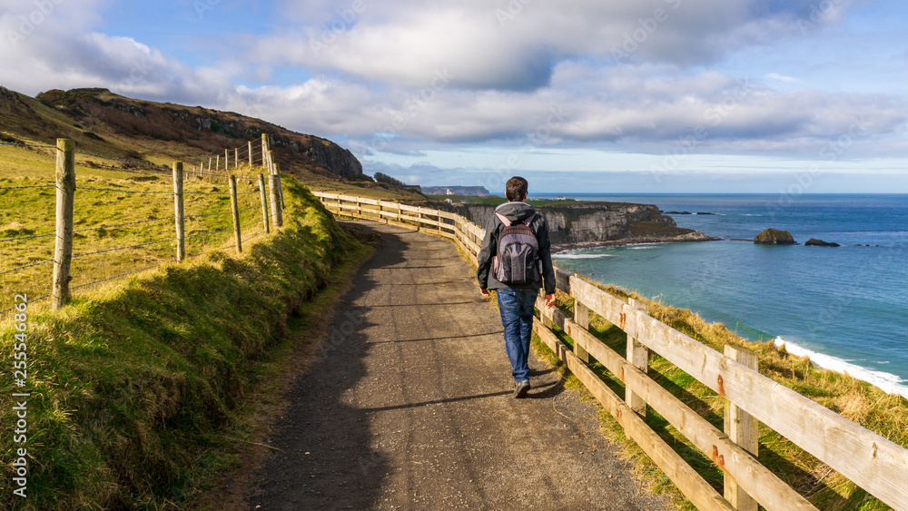 Male Caucasian tourist with a backpack hiking on a trail in a spectacular coastal landscape with sheer cliffs and grass shinnying under the winter sun. Famous Antrim coastline, Northern Ireland.