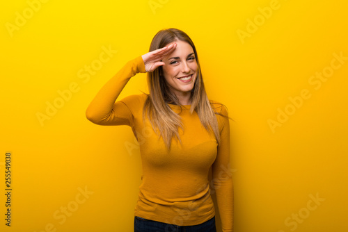 Young woman on yellow background saluting with hand
