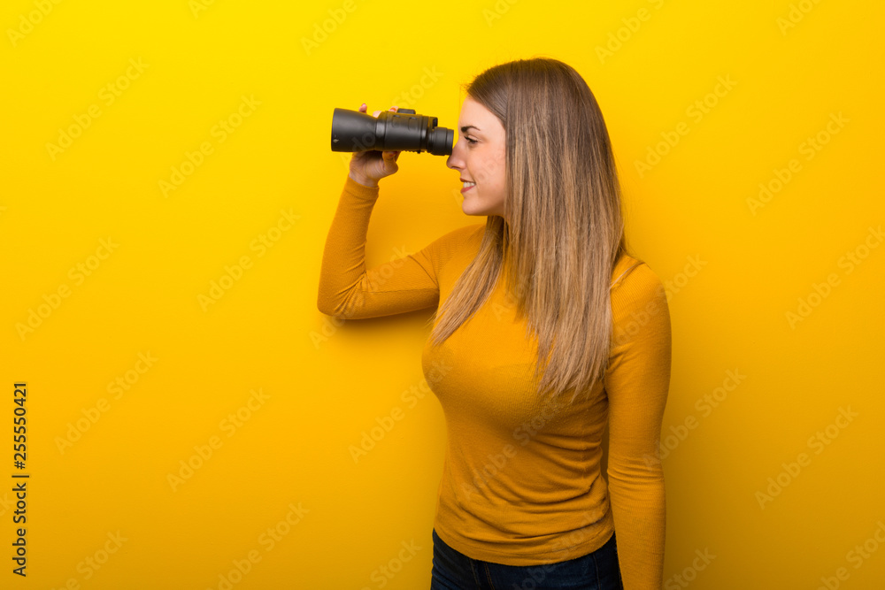 Young woman on yellow background and looking in the distance with binoculars Young woman on yellow background and looking in the distance with binoculars